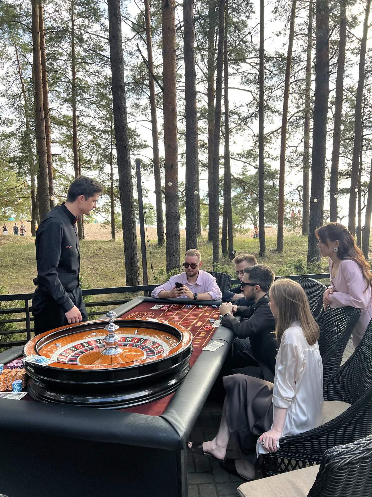Крупье ведёт игру на террасе в сосновом лесу, гости следят за ставками